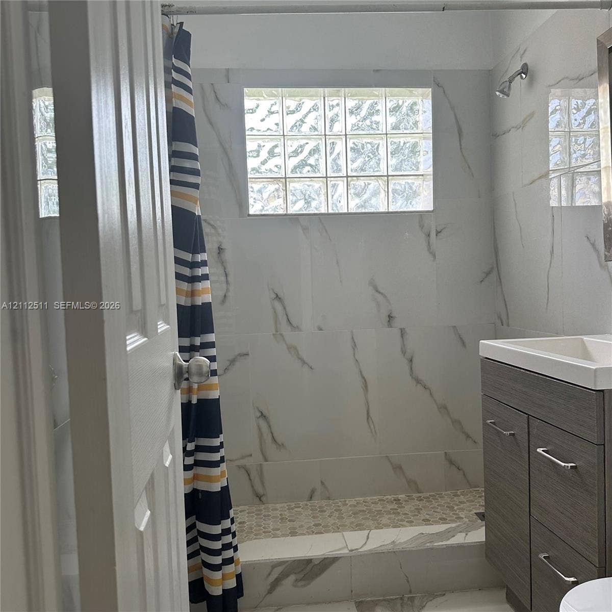 Bathroom featuring large-format wall tiles with veined detailing, glass block window, stone-look pebble tile shower floor, and dark wood-finish vanity with a rectangular integrated sink