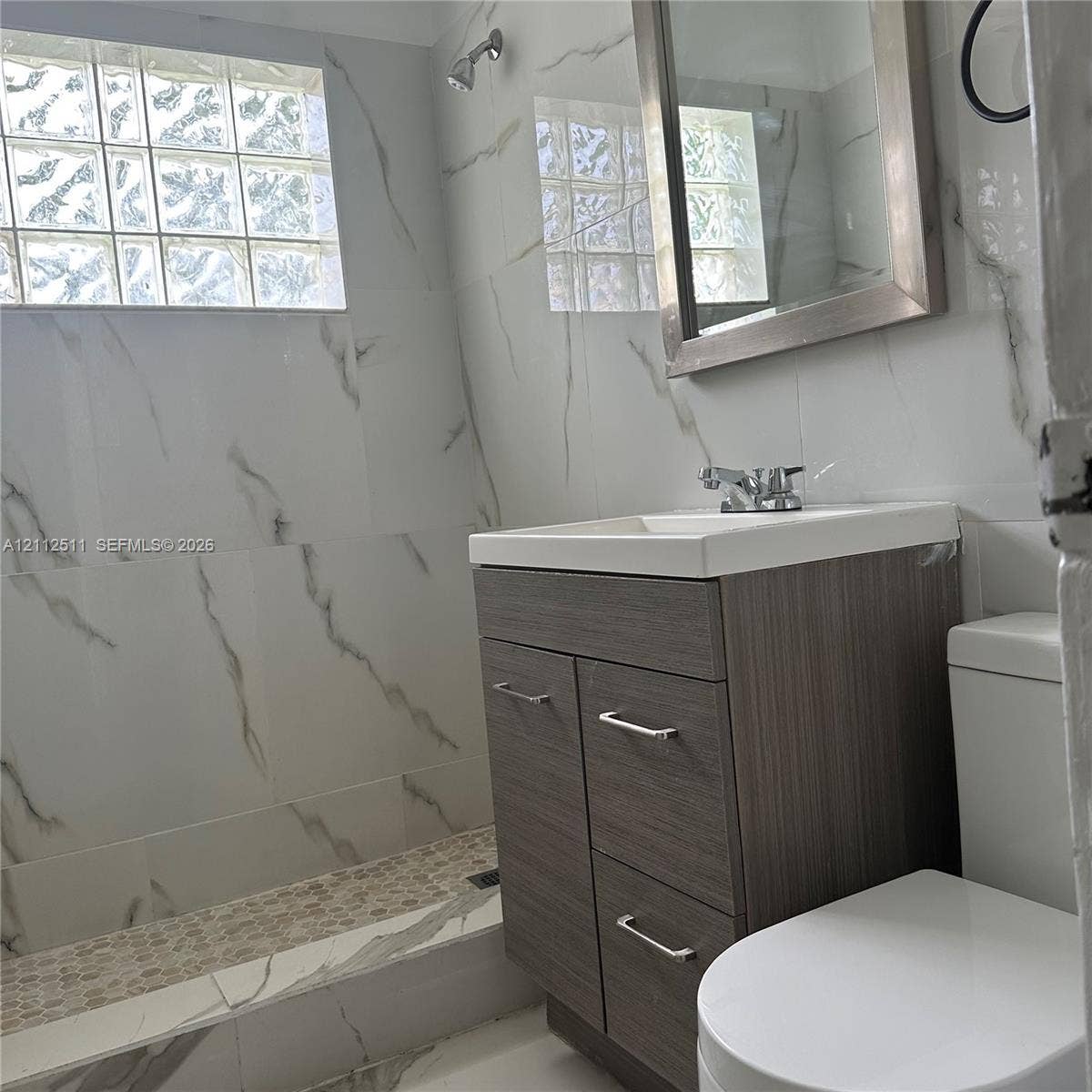 Bathroom featuring large-format stone-patterned wall tile, walk-in shower with pebble-stone flooring, glass block window, dark wood-finish vanity cabinet, and rectangular sink