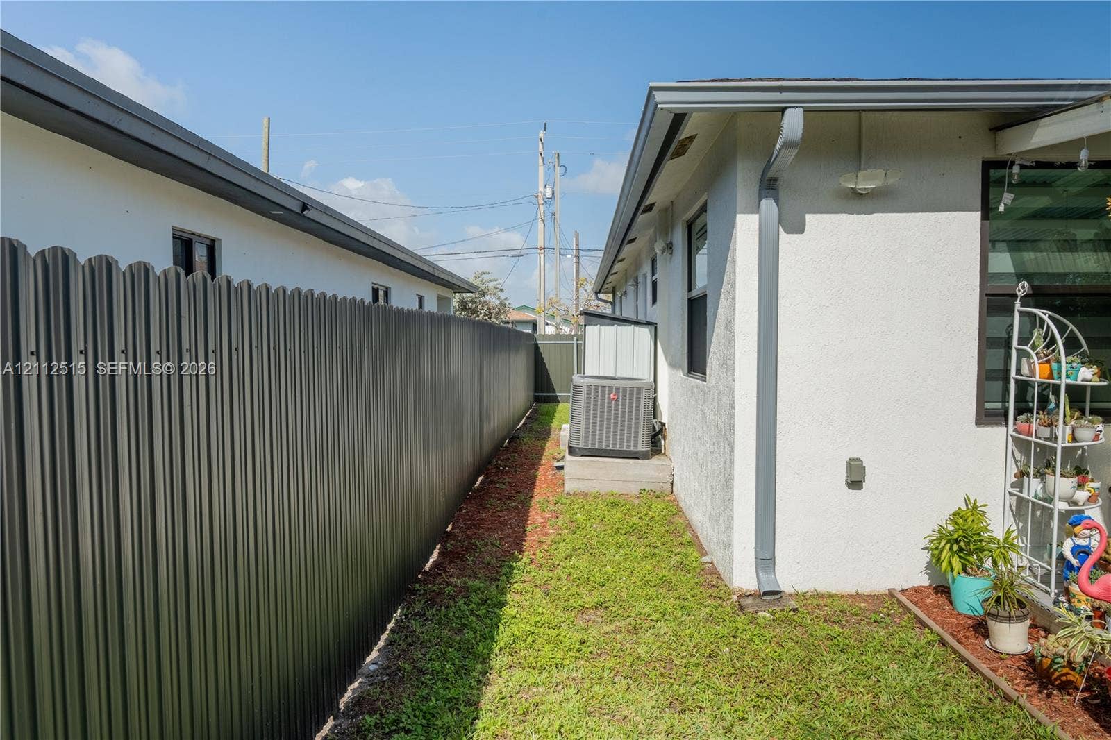 Hallandale Beach, FL 33009 Stucco exterior side yard featuring a corrugated metal privacy fence, aluminum downspout, and exterior HVAC condenser unit on a concrete pad