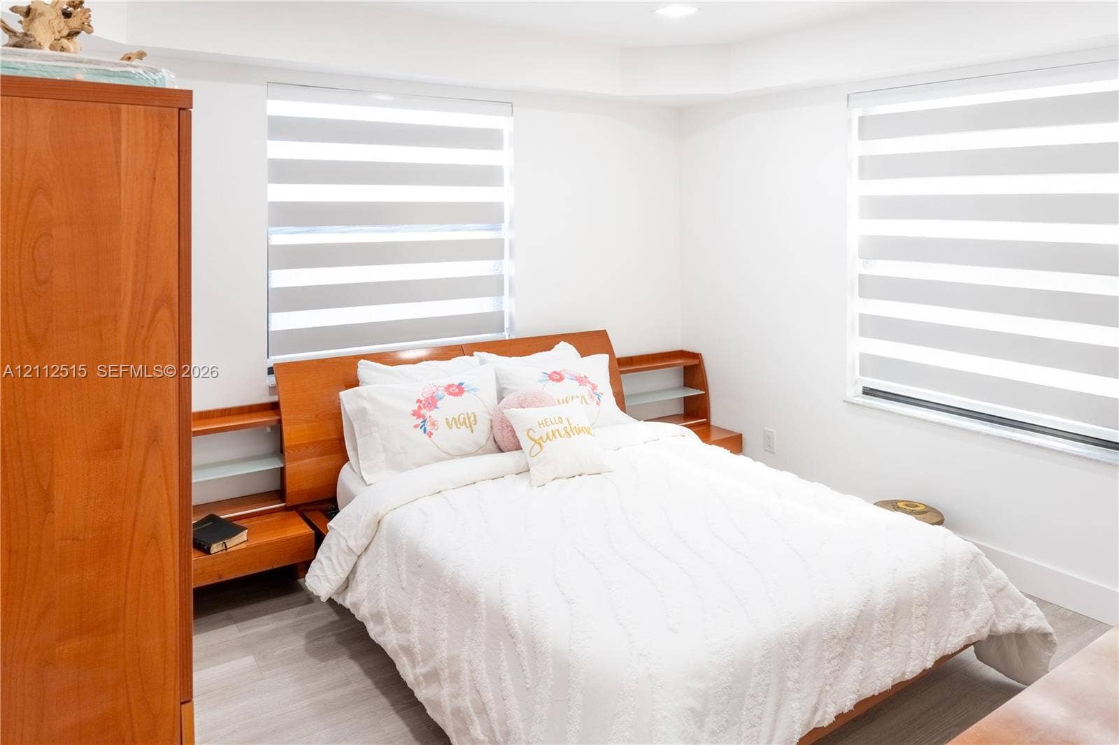 Hallandale Beach, FL 33009 Bedroom featuring wood-finish flooring, dual windows with banded roller shades, and custom built-in wood headboard with integrated shelving
