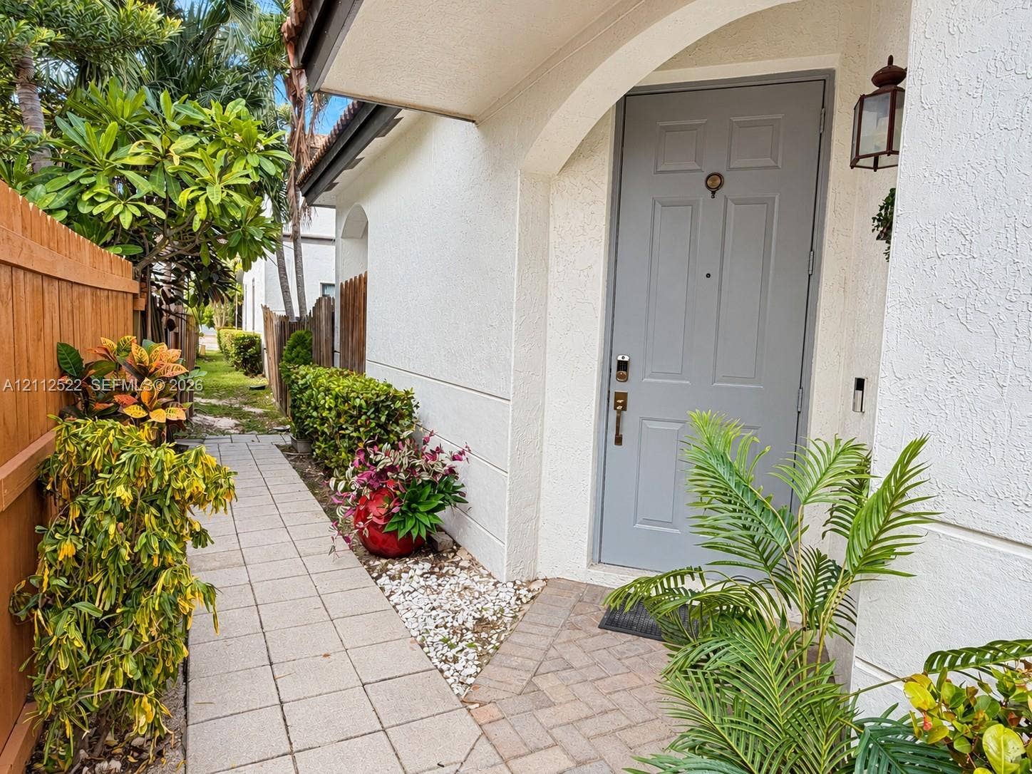 7822 NW 116th Avenue, Doral, FL 33178 Entryway featuring a solid-panel gray front door, stucco exterior finish, arched architectural detail, decorative exterior wall-mounted lantern, and a herringbone-patterned paver landing