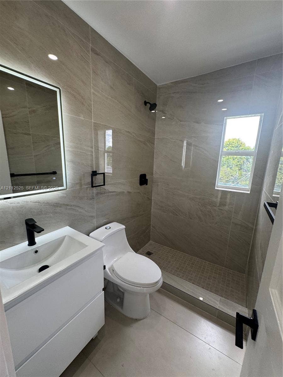 351 E 14th Street, #353, Hialeah, Hialeah, FL 33010 Bathroom featuring wall-to-wall large-format porcelain tile, a floating vanity with a vessel sink, a frameless glass-enclosed walk-in shower, matte black fixtures, and an LED-backlit mirror