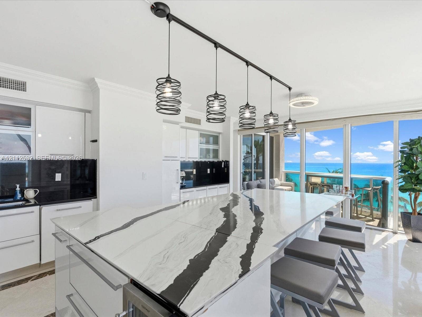 18911 Collins Avenue, #601, Sunny Isles Beach, FL 33160 Kitchen featuring a large central island with veined stone countertop, high-gloss white cabinetry, black tile backsplash, and linear pendant lighting, framed by floor-to-ceiling glass doors with ocean scenery