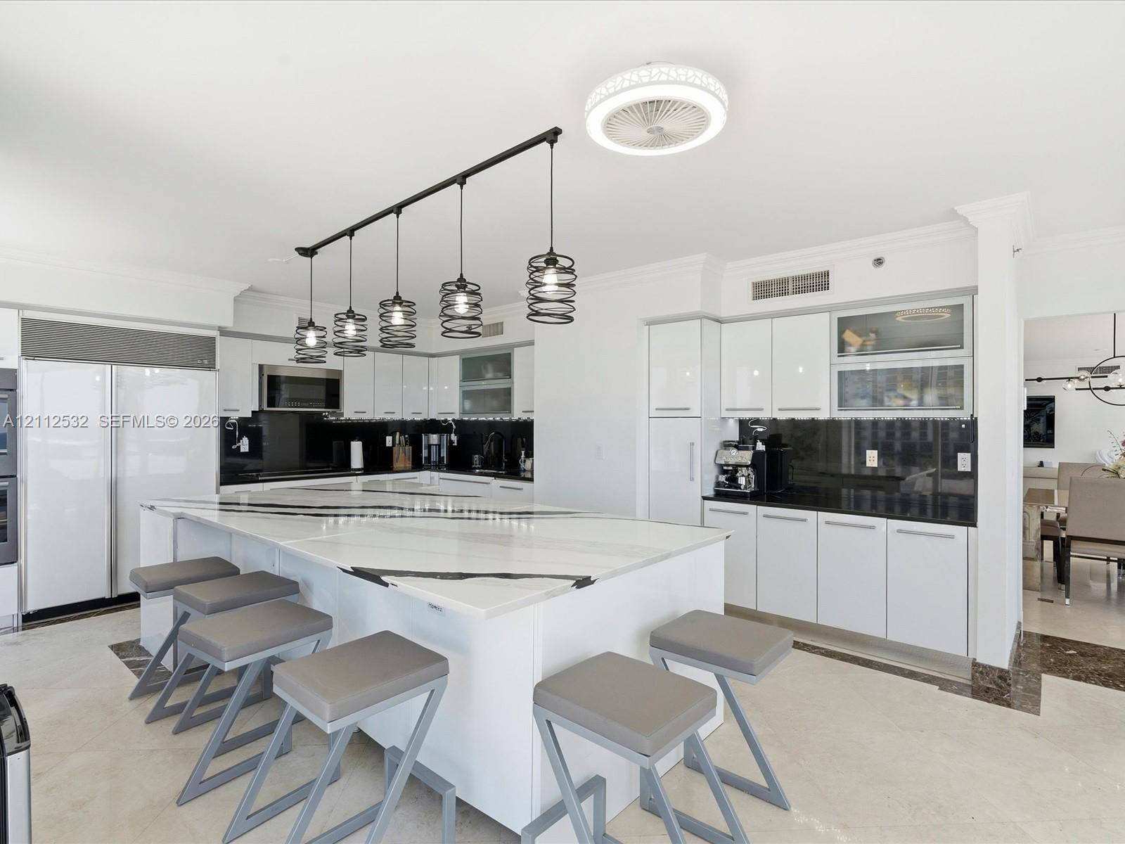18911 Collins Avenue, #601, Sunny Isles Beach, FL 33160 Kitchen featuring a large waterfall-edge island, high-gloss white cabinetry, contrasting dark stone countertops, integrated stainless steel appliances, and linear industrial-style pendant lighting