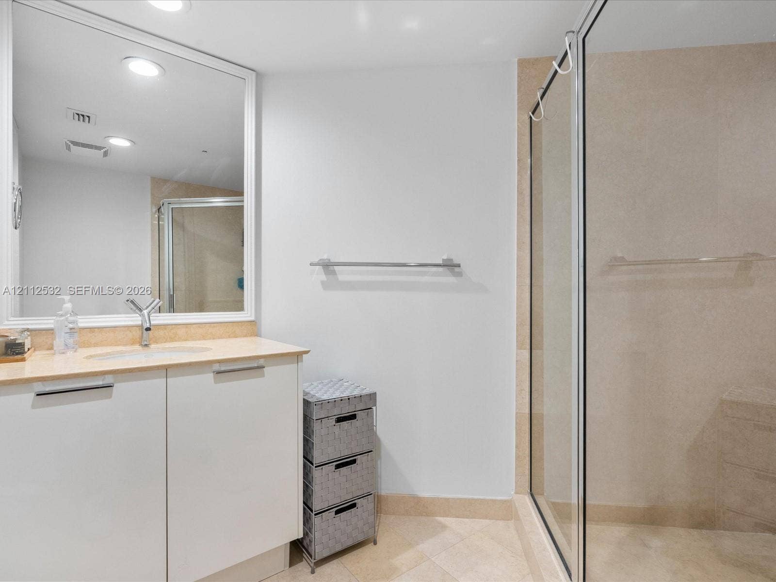 18911 Collins Avenue, #601, Sunny Isles Beach, FL 33160 Bathroom vanity with a light-toned countertop, integrated sink, and white cabinetry, featuring a large framed mirror, stone-tile flooring, and a walk-in shower enclosure with a glass door