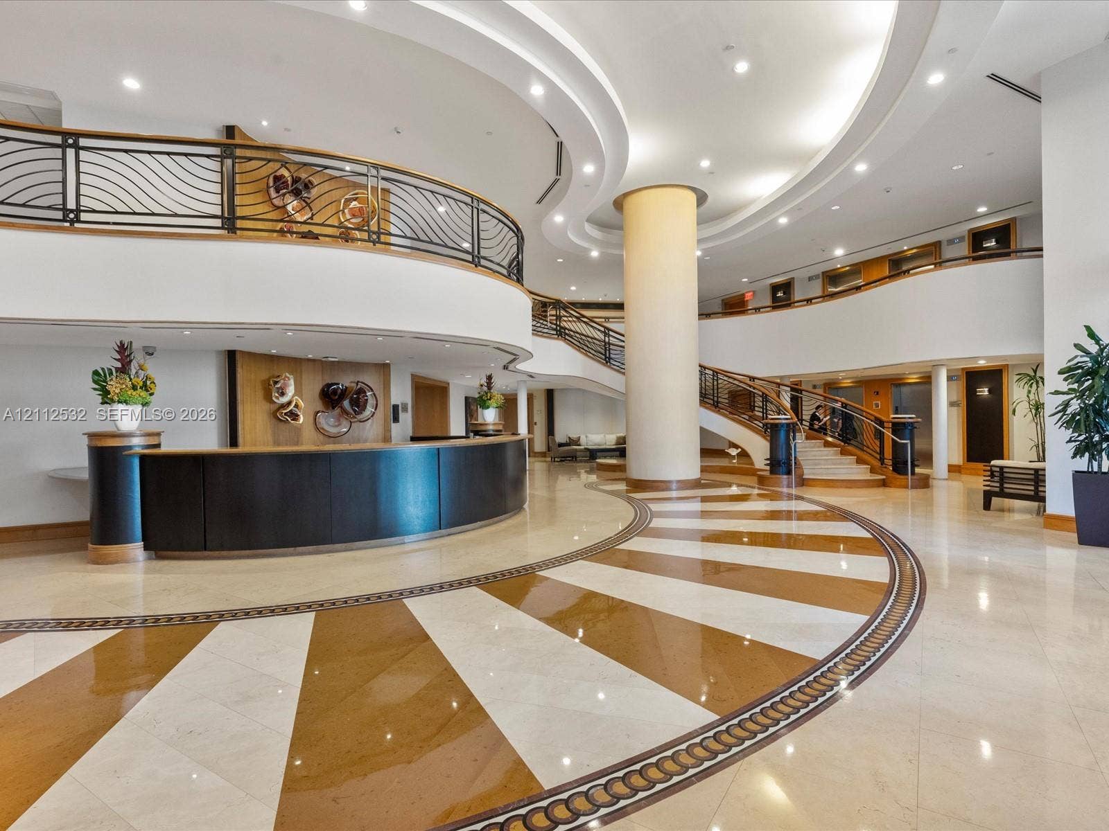 18911 Collins Avenue, #601, Sunny Isles Beach, FL 33160 Grand lobby featuring expansive radial-patterned stone flooring, a curved reception desk with wood paneling, a sweeping staircase with ornate metal railings, and multi-level mezzanine architecture