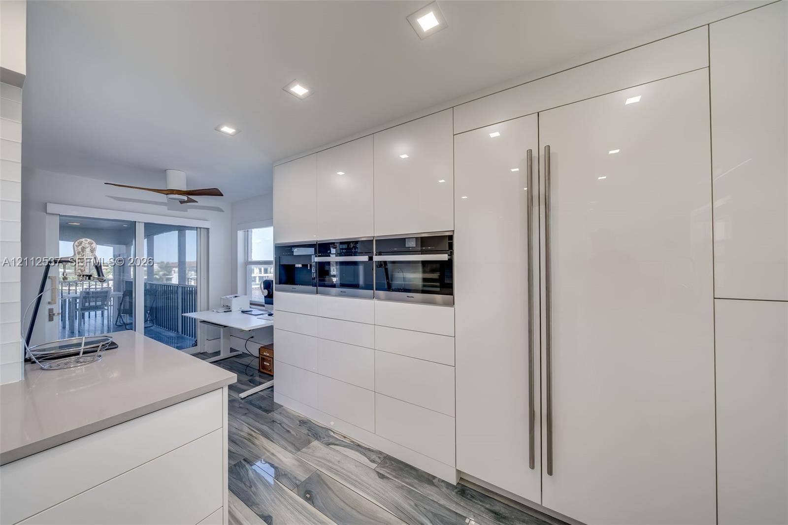 2871 N Ocean Boulevard, #R360, Boca Raton, FL 33431 Sleek, high-gloss white cabinetry featuring integrated, full-height refrigerator and flush-mount wall ovens, quartz countertops, recessed ceiling lighting, and polished stone-finish flooring