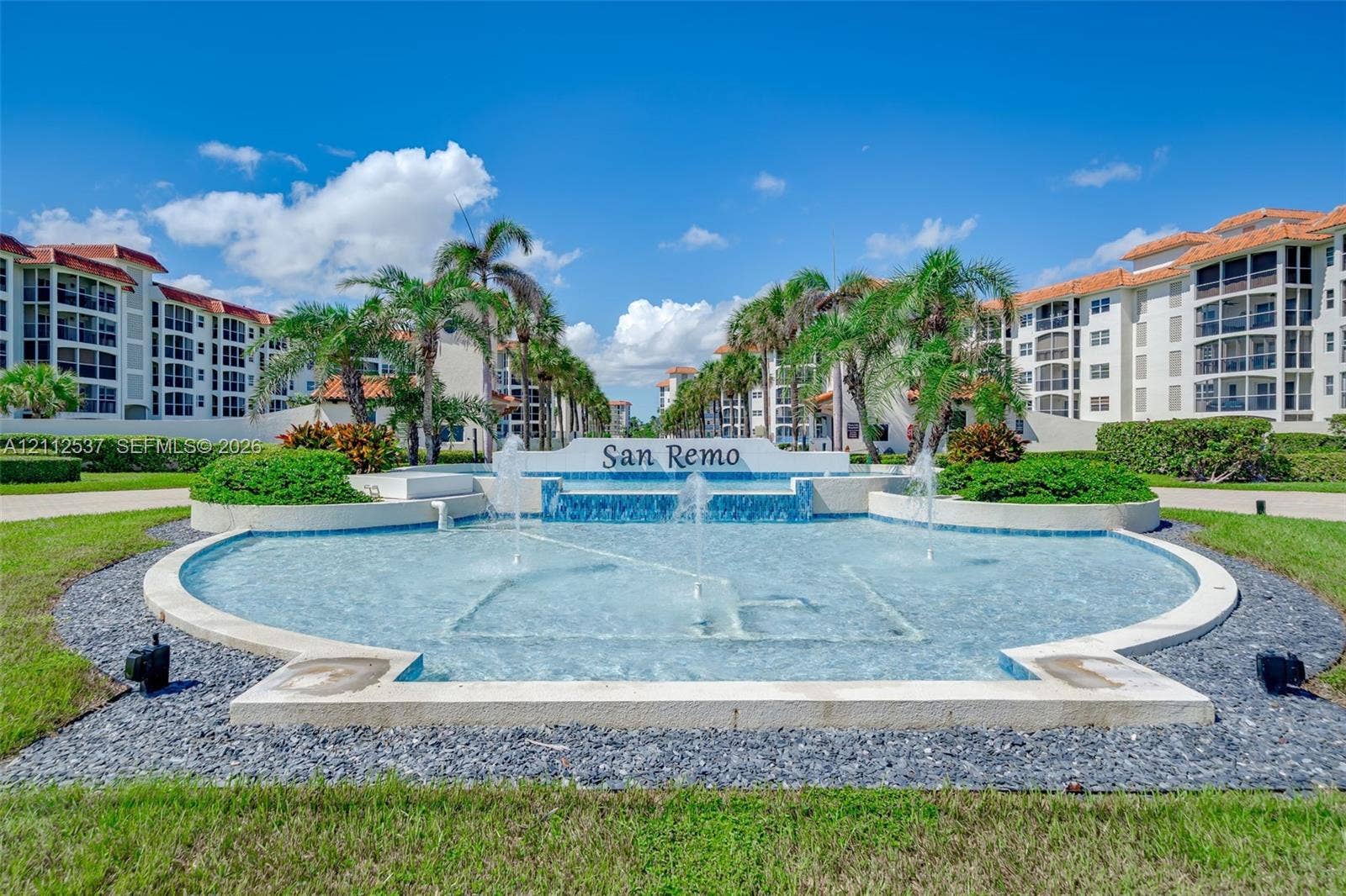 2871 N Ocean Boulevard, #R360, Boca Raton, FL 33431 Gated community entrance featuring a tiered water fountain, mosaic tile accents, mature palm trees, and manicured landscaping