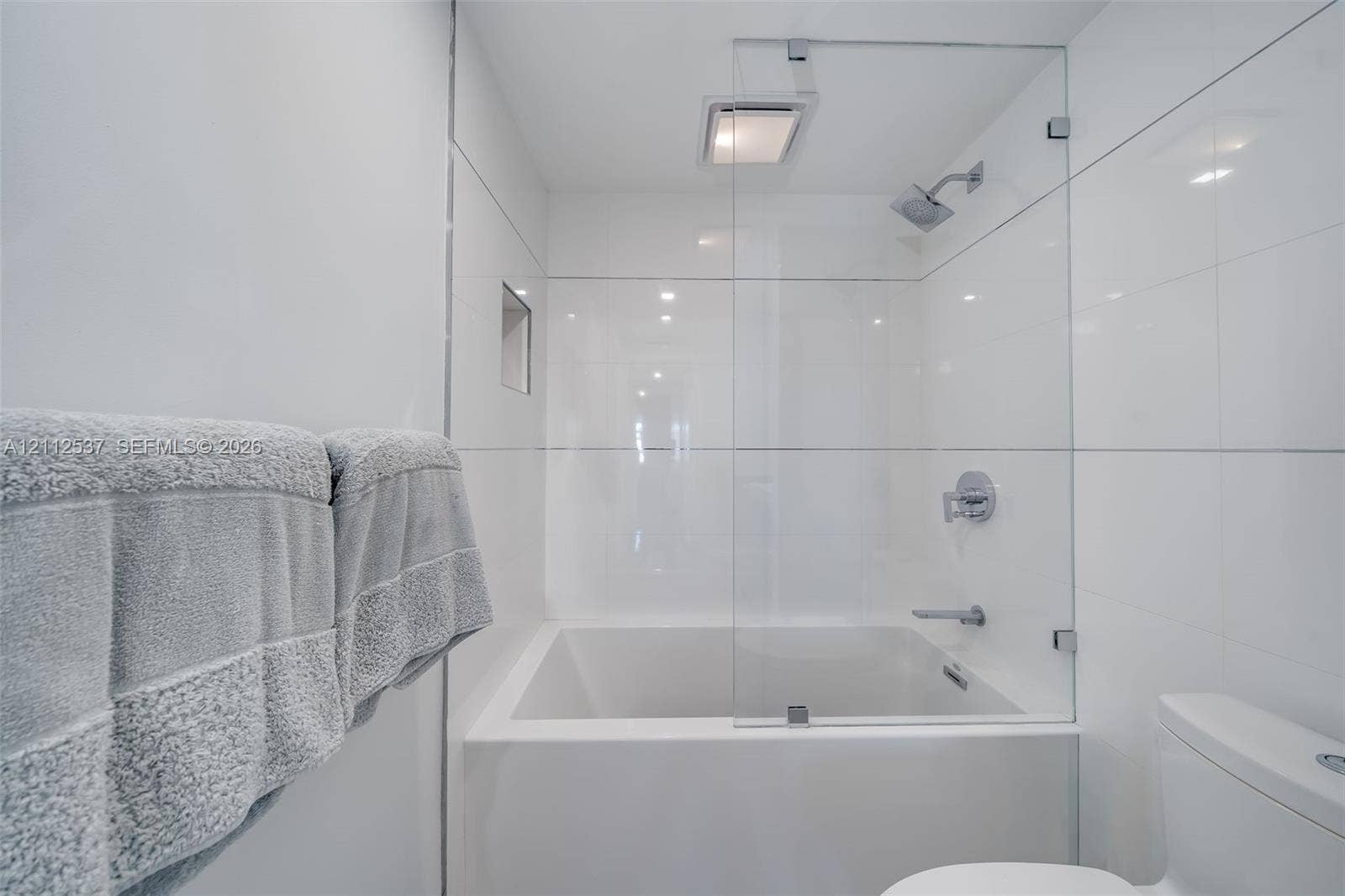 2871 N Ocean Boulevard, #R360, Boca Raton, FL 33431 Soaking tub with a frameless glass splash panel, chrome rainfall showerhead, recessed wall niche, and floor-to-ceiling white tile surround