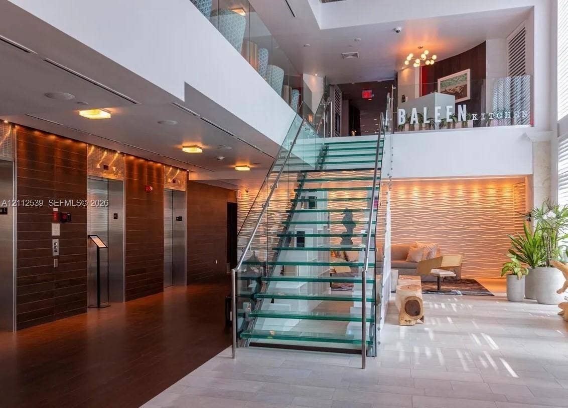 17315 Collins Avenue, #705, Sunny Isles Beach, FL 33160 Grand lobby entrance featuring a central glass-tread staircase with stainless steel railings, multi-level design, wood-paneled elevator surround, and textured wall accents