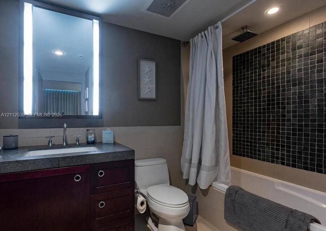 17315 Collins Avenue, #705, Sunny Isles Beach, FL 33160 Bathroom featuring a dark wood-finish vanity with a stone countertop, an illuminated mirror, and a bathtub with black mosaic tile surround and rainfall shower head