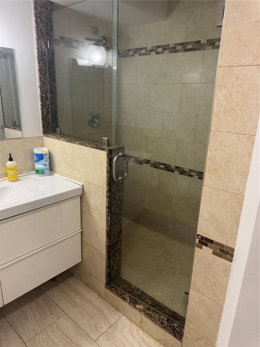 1301 NE Miami Gardens Drive, #515W, Miami, Miami, FL 33179 Walk-in shower with a glass door, stone-surround accents, large-format wall tiling with decorative inlay, and a white wall-mounted vanity