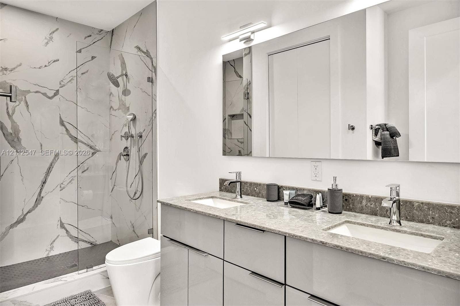 Bathroom featuring a dual-sink vanity with stone countertop, walk-in shower with floor-to-ceiling tile surround, glass partition, and wall-mounted light fixture