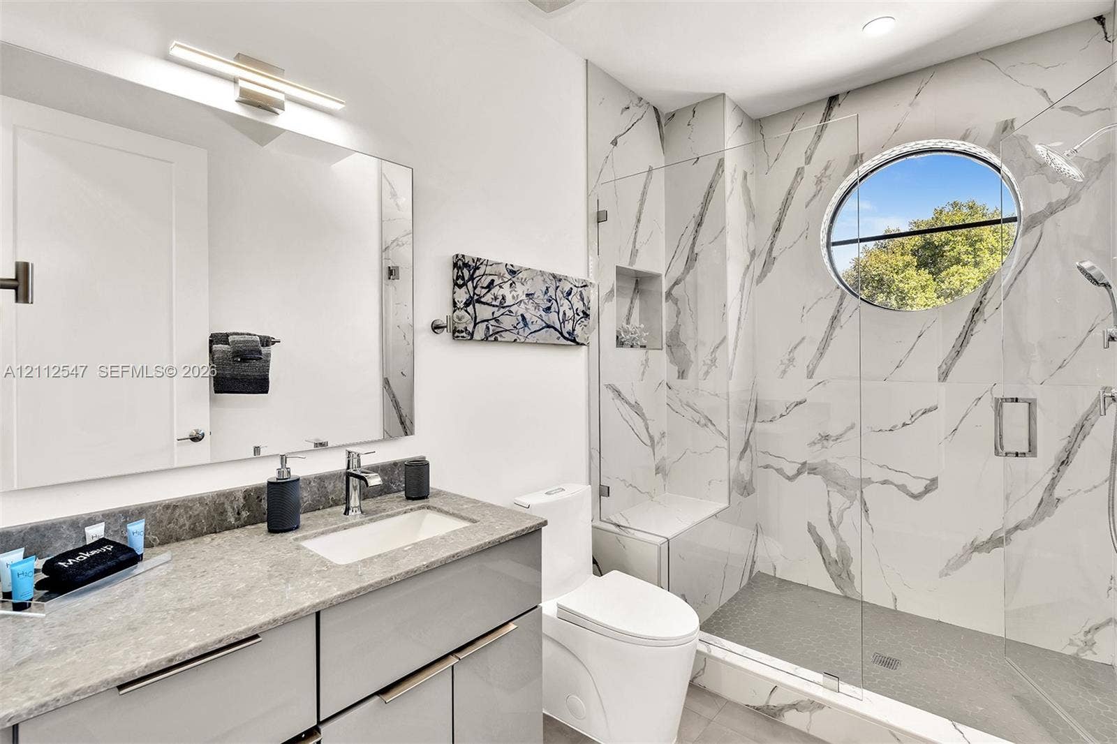 Bathroom featuring a sleek vanity with a stone countertop, an integrated rectangular sink, wall-to-wall mirror, walk-in shower with floor-to-ceiling stone-patterned wall tile, and a decorative circular window