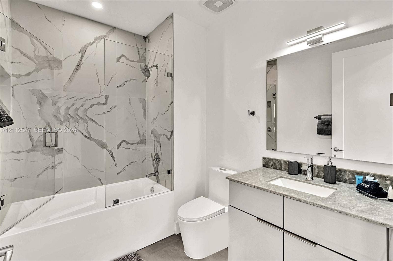 Bathroom featuring a tub-shower combination with floor-to-ceiling large-format tile surround, a glass shower panel, a floating vanity with a stone countertop and rectangular undermount sink, and an expansive wall-mounted mirror