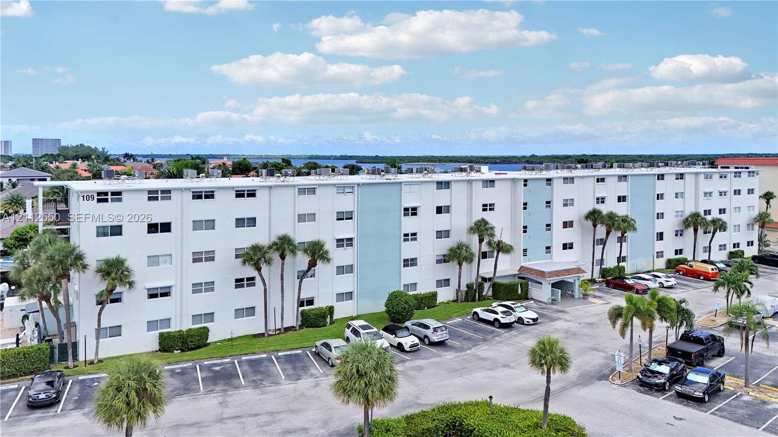 Multi-story residential building exterior with a light-toned stucco facade, ground-level parking area, and mature palm landscaping