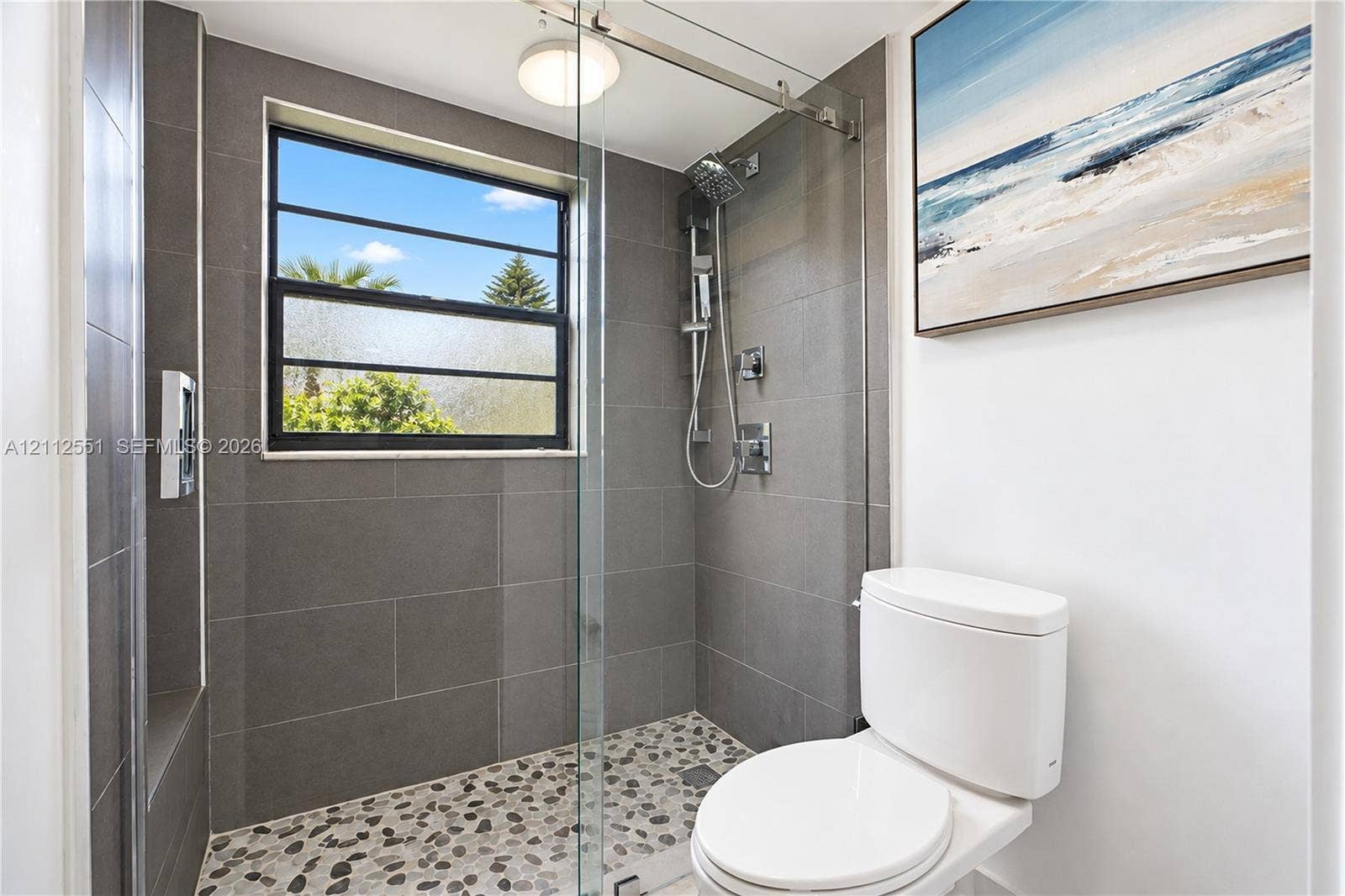 Walk-in shower featuring large-format grey wall tile, a pebble-stone floor, glass panel enclosure, and a chrome rainfall shower head