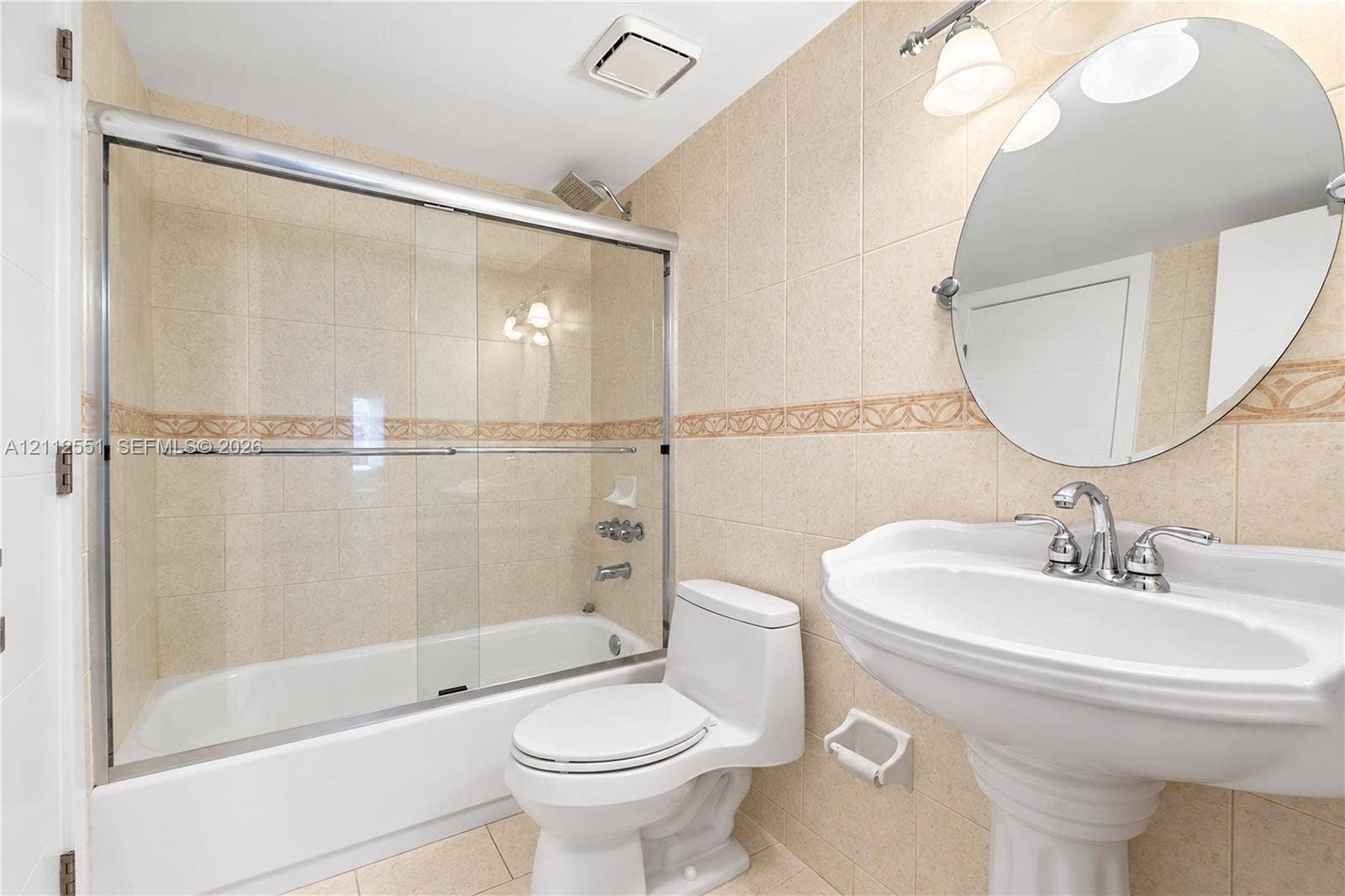 Bathroom featuring a pedestal sink with a chrome faucet, oval wall-mounted mirror, ceramic tile walls with decorative inlay, and a tub-shower combination with a glass sliding enclosure