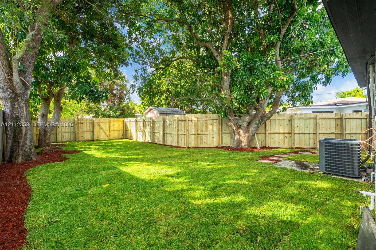 Fenced backyard featuring mature trees, manicured lawn, and organic mulch ground cover