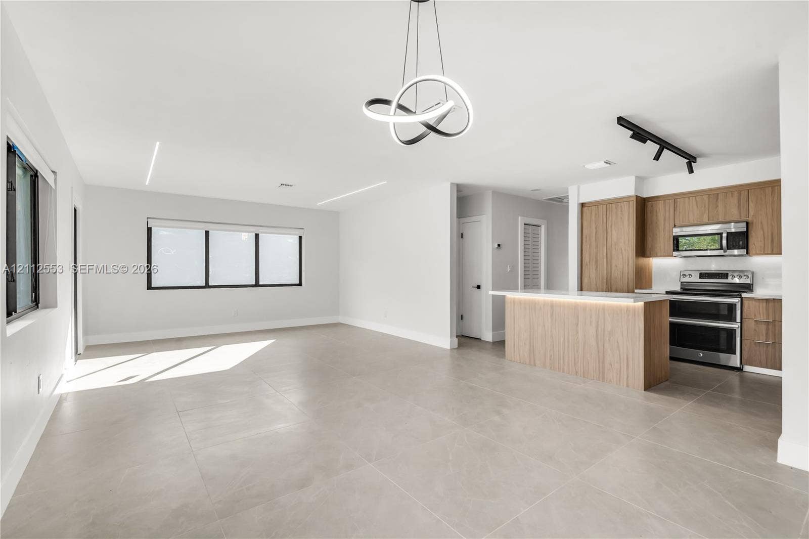 Open-concept space featuring large-format tile flooring, modern circular chandelier, recessed linear lighting, and a kitchen equipped with wood-finish cabinetry, quartz countertops, and stainless steel appliances