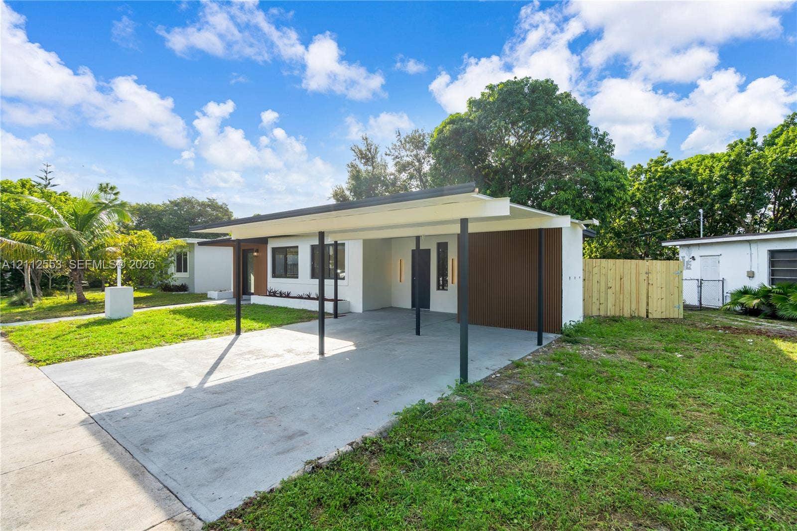 Mid-century inspired exterior featuring a concrete carport, dark wood-tone siding accents, clean-lined vertical wall sconces, and a partial wood privacy fence