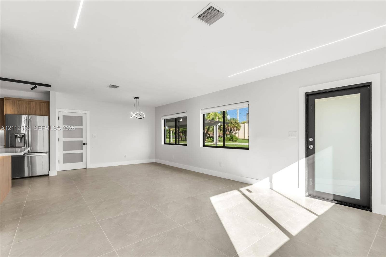 Open-concept interior featuring large-format tile flooring, modern linear recessed lighting, a stainless steel refrigerator, and a contemporary entry door with a glass pane