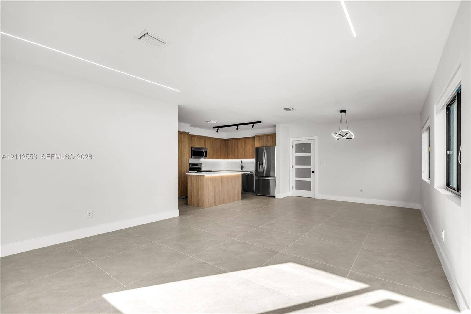 Open-concept interior featuring large-format tile flooring, recessed linear lighting, kitchen with wood-finish cabinetry, center island, and a multi-pane frosted glass door