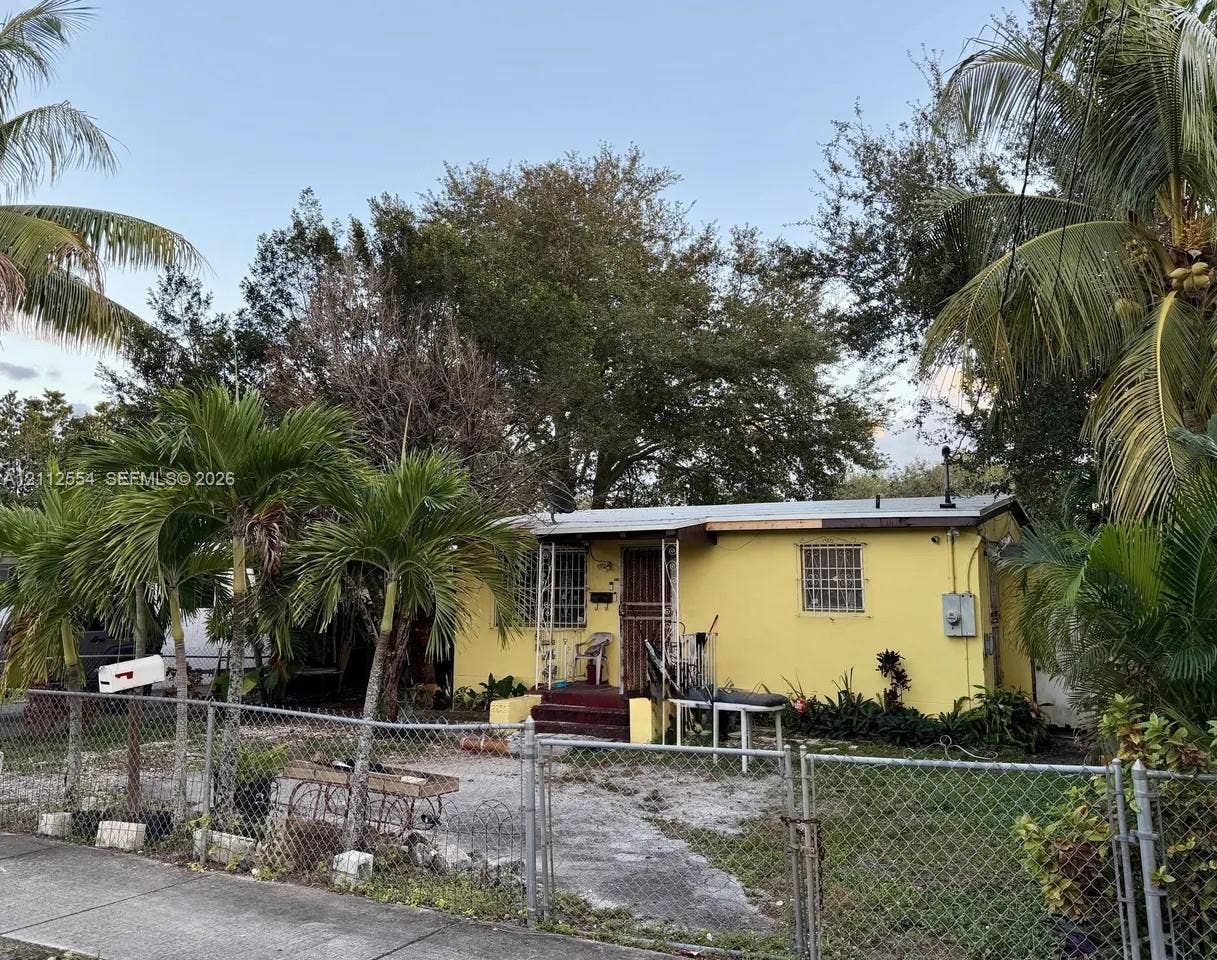 Single-story residence featuring a painted stucco exterior, metal-framed window guards, covered front entryway, and mature palm landscaping situated within a chain-link fenced perimeter