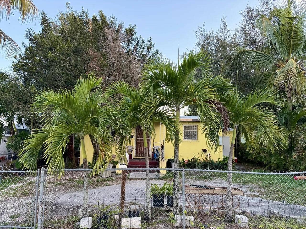 Single-story exterior featuring a stucco facade, mature palm trees, and a chain-link perimeter fence
