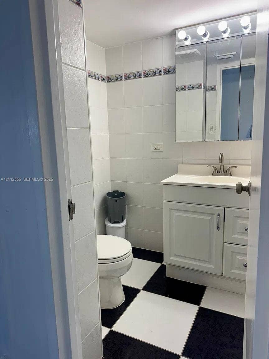 8400 Byron Avenue, #4E, Miami Beach, Miami Beach, FL 33141 Bathroom featuring black-and-white checkered tile flooring, white wall tiling with decorative border, vanity cabinet with integrated sink, and mirrored medicine cabinet with overhead lighting