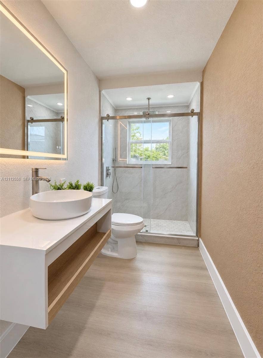 Floating vanity with an integrated vessel sink, illuminated wall mirror, glass-enclosed shower featuring large-format tile surround, and light-toned wood-finish flooring