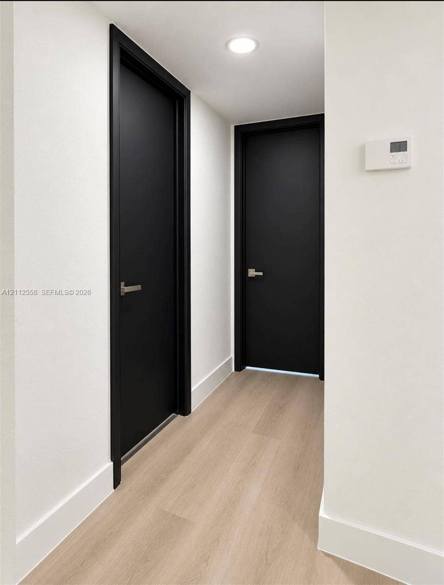 Hallway featuring light-toned wood-finish flooring, solid matte black doors with minimalist metal lever handles, recessed ceiling lighting, and smooth white walls