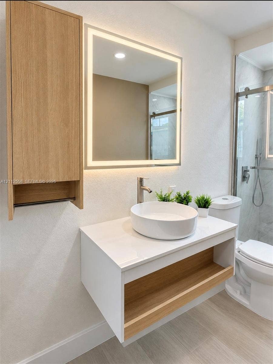Floating vanity with a white countertop and wood-finish open shelving, circular vessel sink, wall-mounted LED-backlit mirror, matching wood-finish wall cabinet, and walk-in shower with a glass enclosure