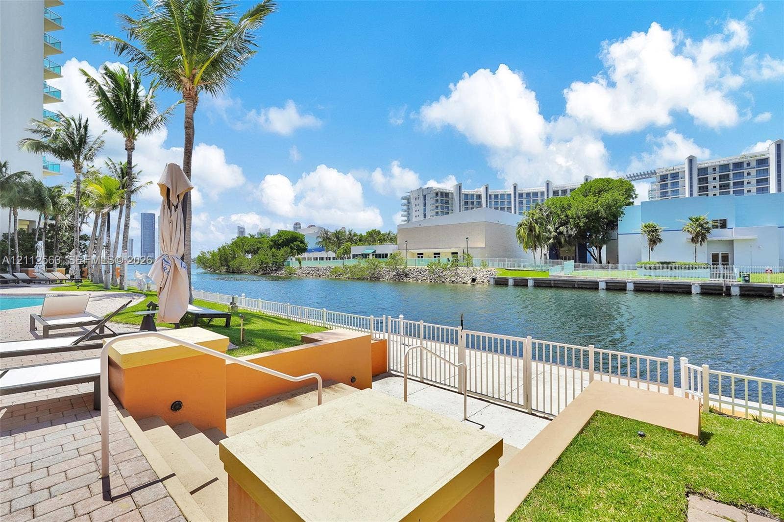 3330 NE 190th Street, #2016, Aventura, FL 33180 Waterfront patio area featuring paved stone decking, built-in tiered retaining walls, a metal safety railing, and mature palm trees