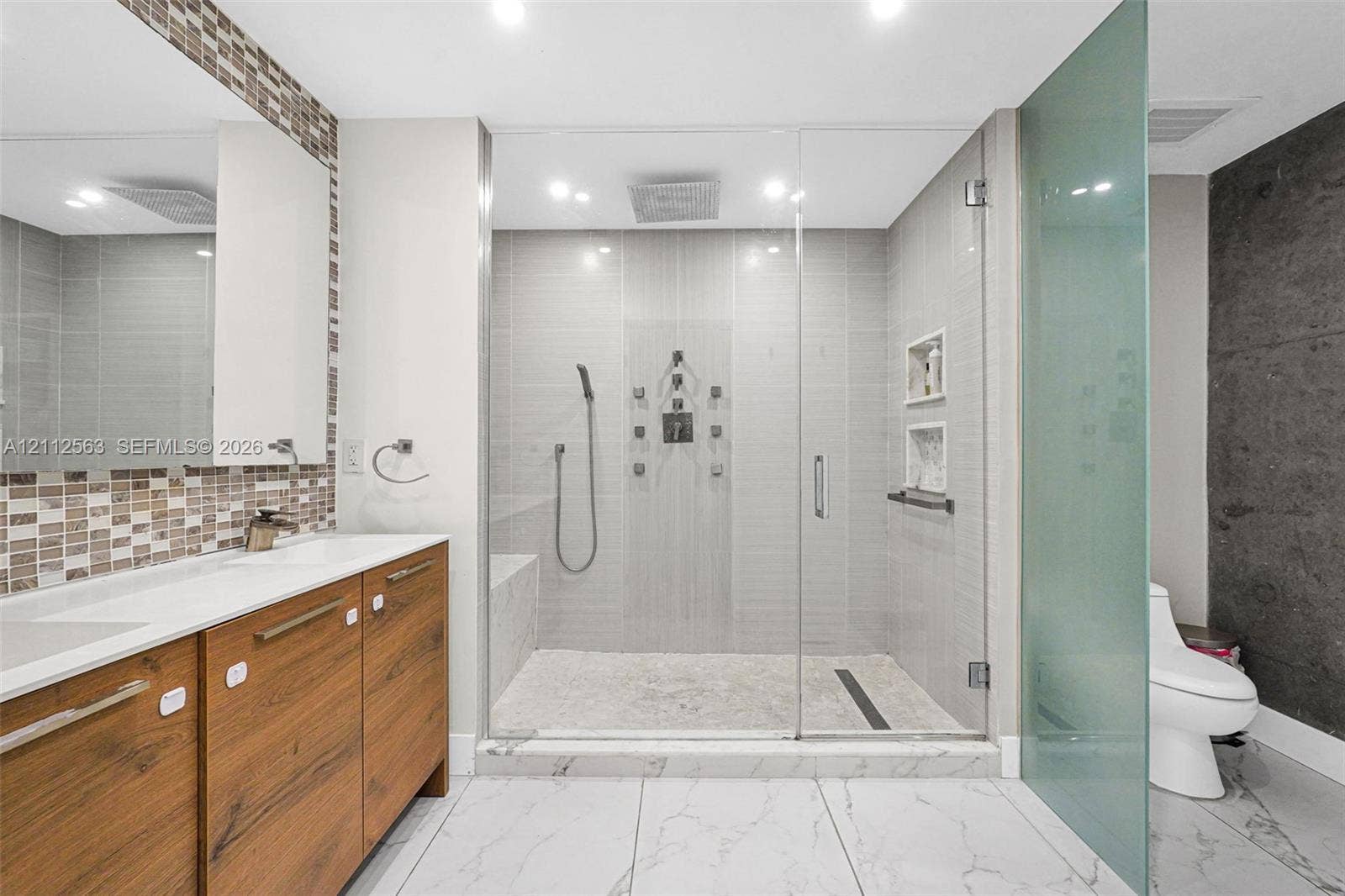 3330 NE 190th Street, #2016, Aventura, FL 33180 Bathroom featuring a floating wood-finish double vanity, walk-in glass-enclosed shower with multiple spray heads, built-in shelving, stone-finish flooring, and mosaic tile backsplash