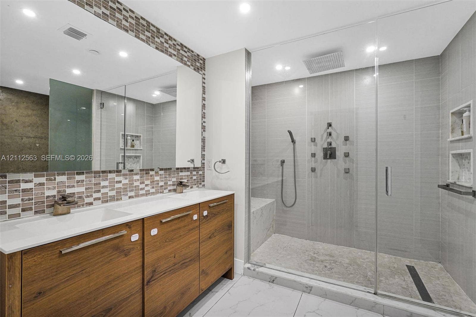 3330 NE 190th Street, #2016, Aventura, FL 33180 Double vanity featuring wood-finish cabinetry, solid-surface countertops, and a full-width mirror with mosaic tile accents