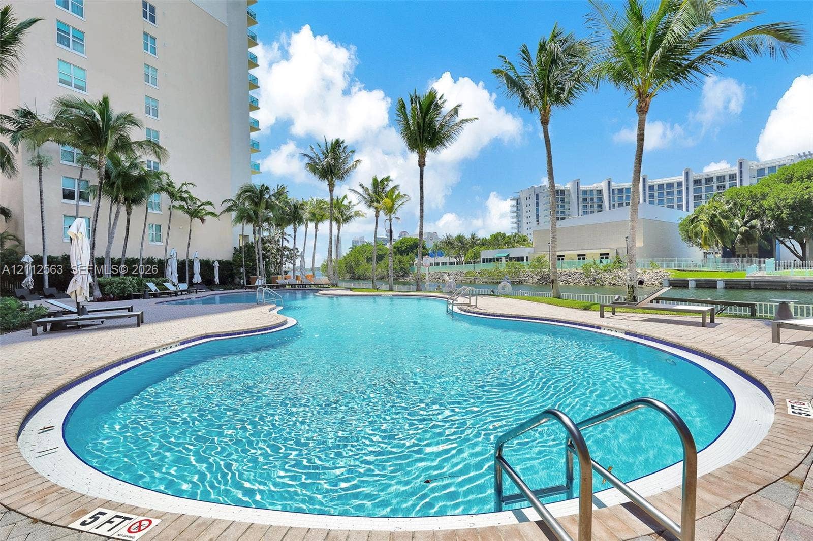 3330 NE 190th Street, #2016, Aventura, FL 33180 Curved swimming pool featuring a brick-paver deck, stainless steel entry rails, and surrounding palm trees