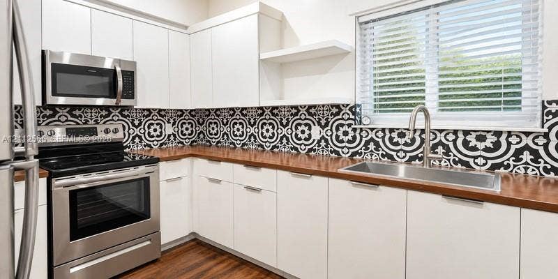 Kitchen featuring white flat-panel cabinetry, wood-finish countertops, stainless steel appliances, a patterned tile backsplash, and a stainless steel undermount sink