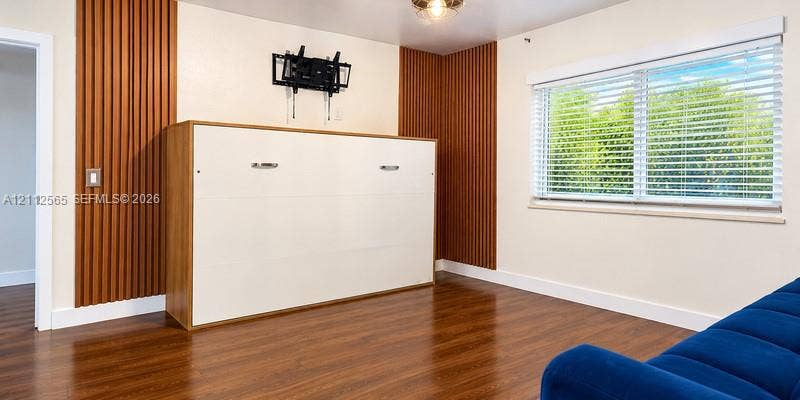 Room featuring wood-finish flooring, vertical wood-slat accent wall detailing, white built-in cabinetry, and a window with horizontal blinds