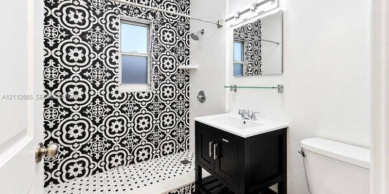 Bathroom featuring a black-and-white patterned tile shower surround, black vanity cabinet with an integrated white sink, rectangular wall mirror, and overhead vanity lighting