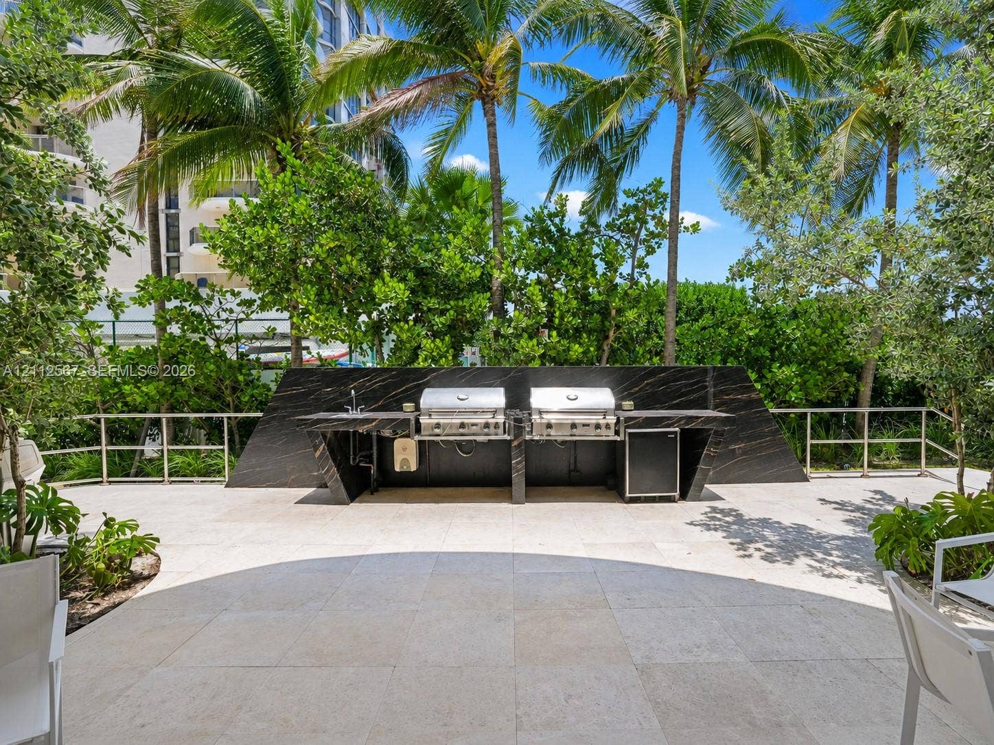 6365 Collins Avenue, #3208, Miami Beach, Miami Beach, FL 33141 Outdoor kitchen featuring a dark stone-finish backsplash and countertop, stainless steel dual grills, integrated refrigeration, and a stone-tiled patio surrounded by mature palm trees