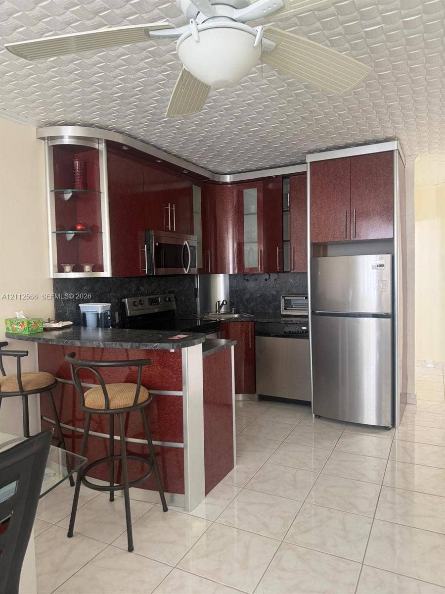 1936 S Ocean Drive, #8B, Hallandale Beach, Hallandale Beach, FL 33009 Kitchen featuring high-gloss cherry-finish cabinetry, stone-texture countertops, integrated stainless steel appliances, a peninsula breakfast bar, and tile flooring