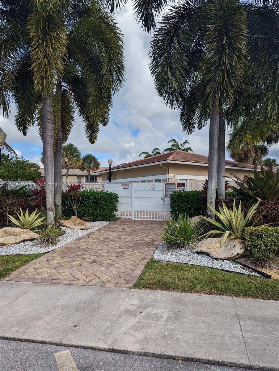 Paver stone walkway leading to a gated entry, accented by mature palm trees, decorative rock borders, and manicured landscaping
