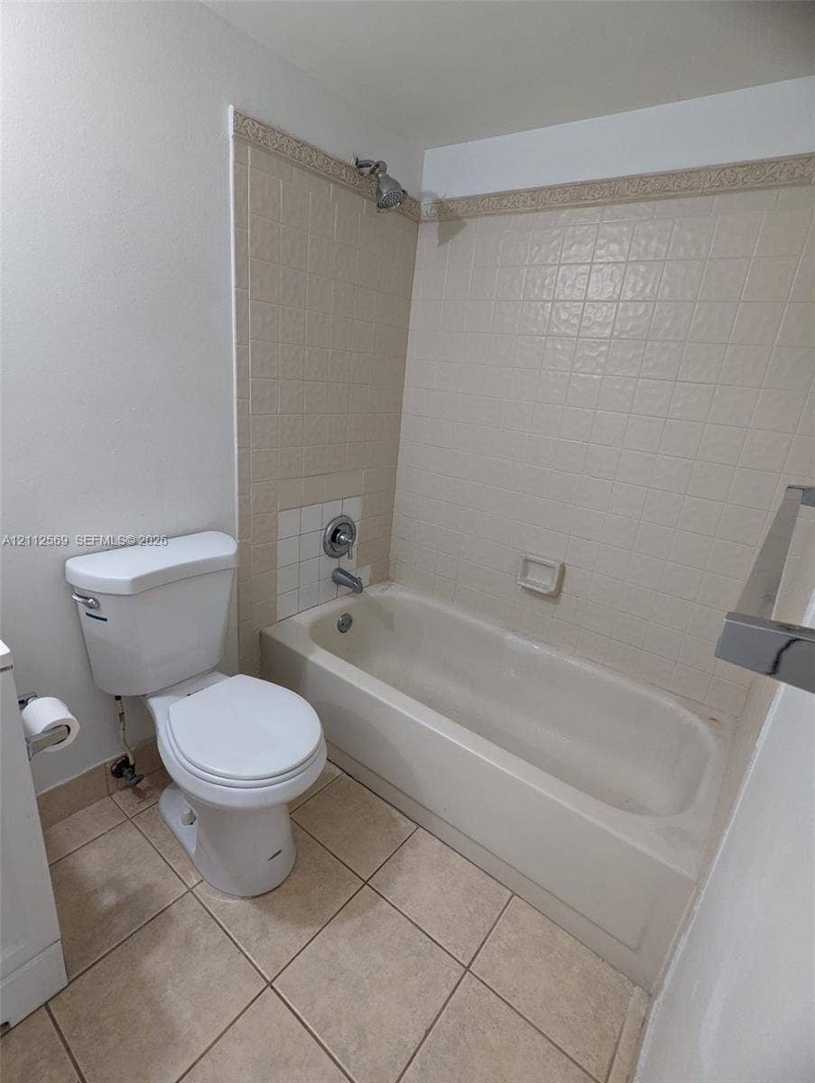 Full bathroom featuring a white porcelain toilet, a standard bathtub with a showerhead, beige square wall tiling with a decorative border, and light-toned tile flooring