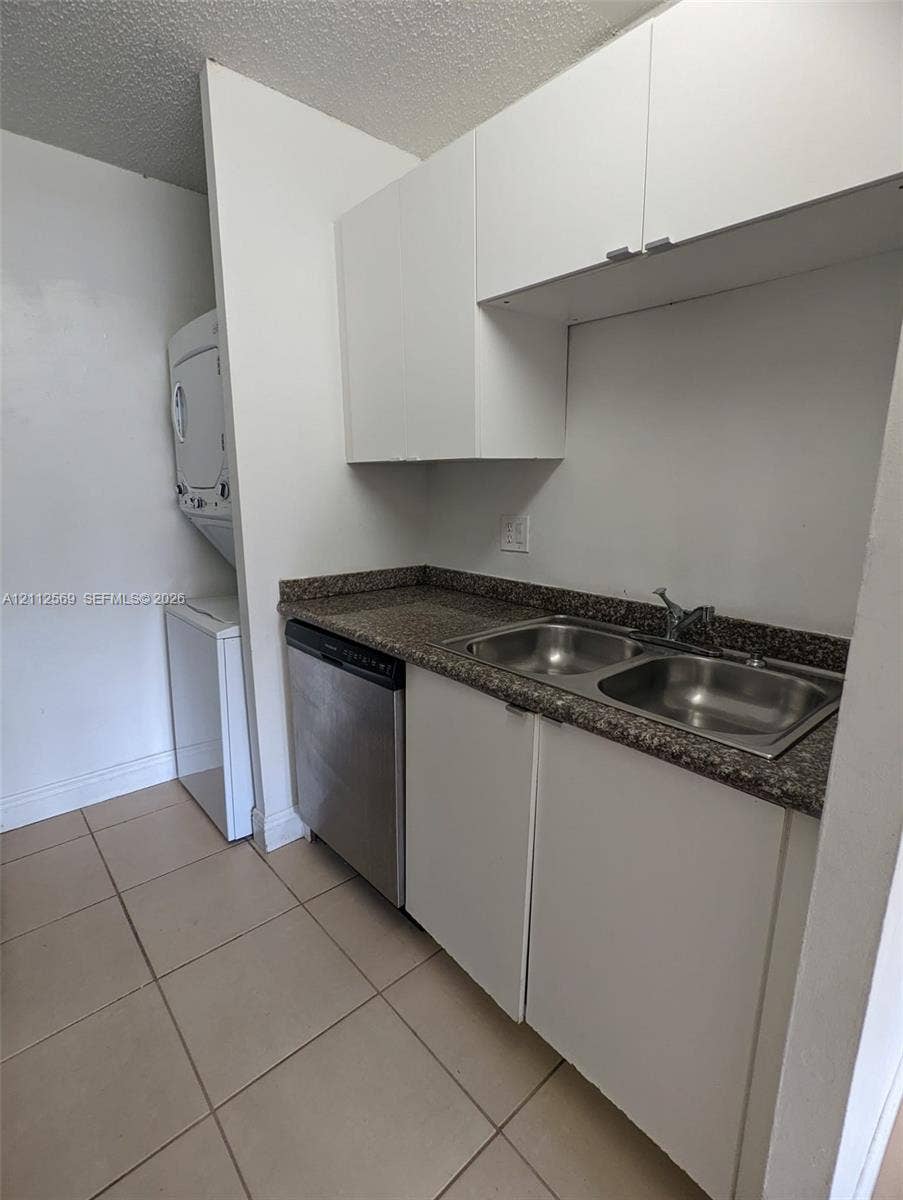 Utility space featuring white cabinetry, a dual-basin stainless steel sink, a stone-finish countertop, a stainless steel dishwasher, and stacked laundry appliances