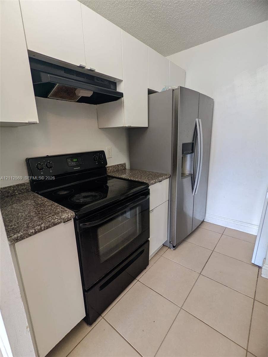 Kitchen featuring white cabinetry, stone-finish countertops, tile flooring, a stainless steel refrigerator, and a black electric range with a built-in exhaust hood
