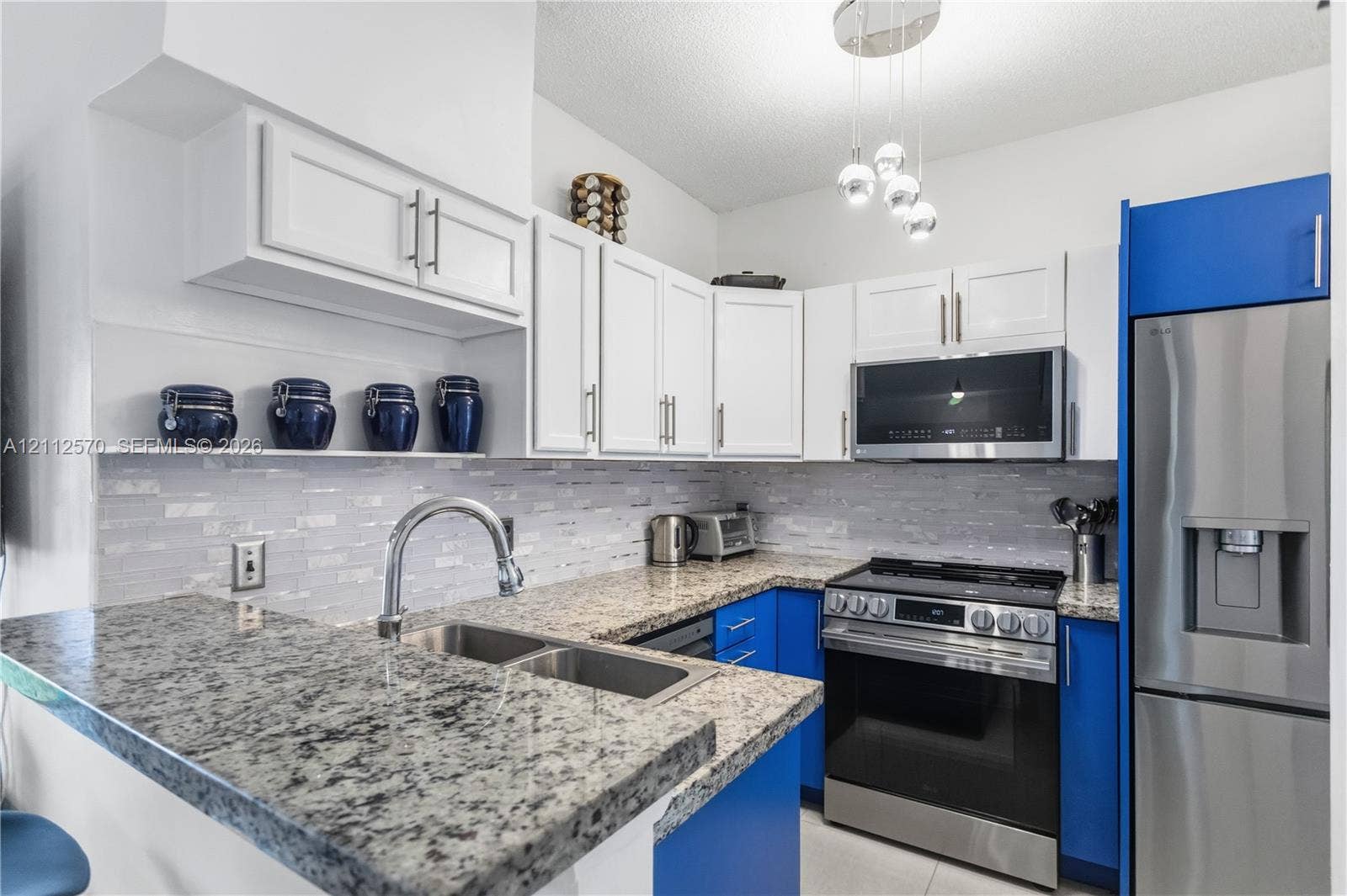 Kitchen featuring two-tone white and blue cabinetry, speckled stone countertops, a multi-textured glass tile backsplash, stainless steel appliances, and a polished metal gooseneck faucet