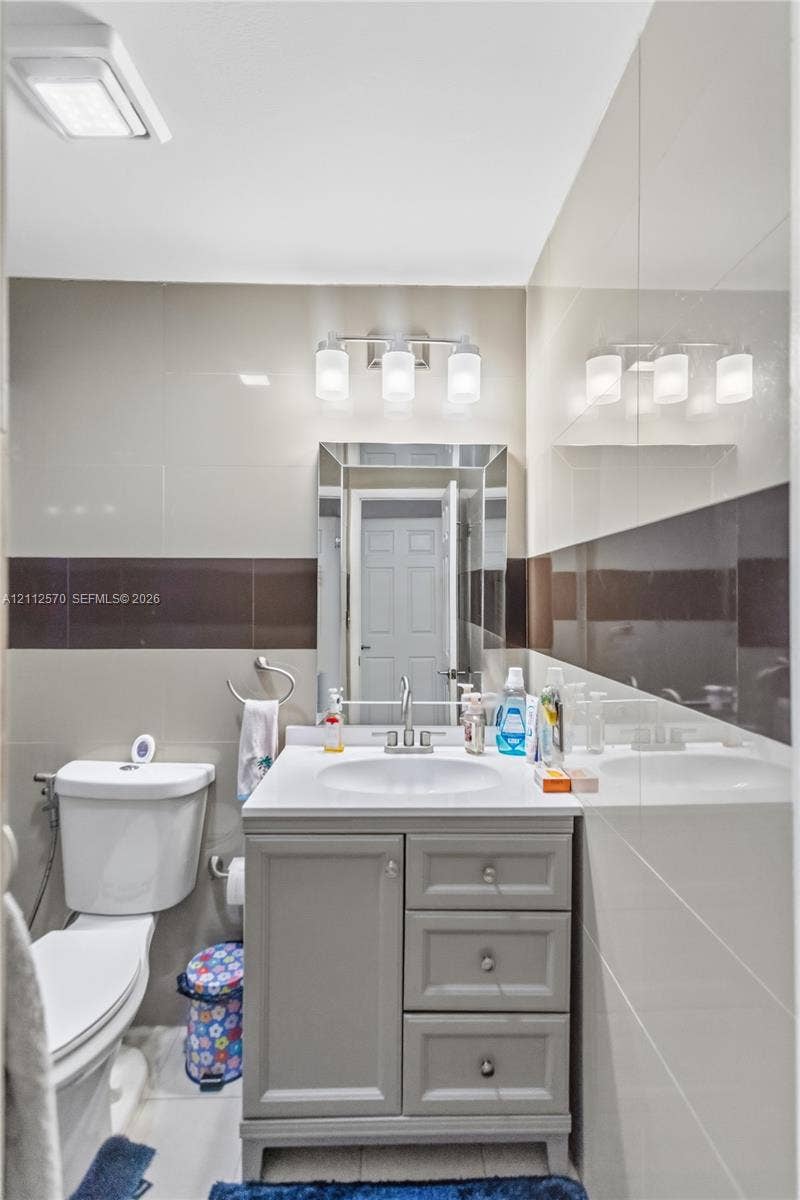 Bathroom featuring a grey vanity with three storage drawers, white countertop with integrated sink, tri-light fixture, framed wall mirror, and large-format wall tile with contrasting horizontal inlay