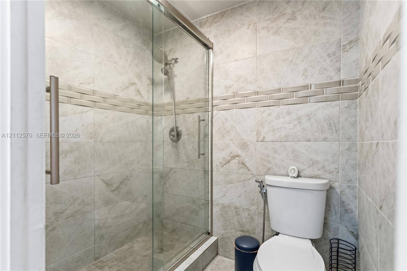 Walk-in shower with a glass enclosure, brushed nickel handheld showerhead, floor-to-ceiling stone-patterned tile, and decorative horizontal tile accent border