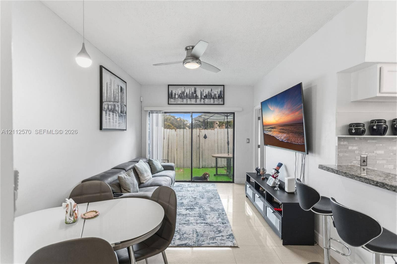 Neutral-toned tile flooring, ceiling-mounted fan with integrated light, sliding glass doors, and a stone-surface breakfast bar